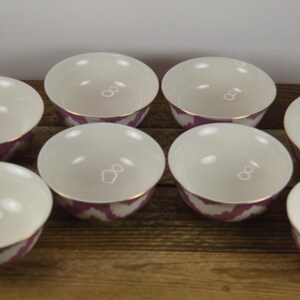 May include: Set of eight white ceramic bowls with a pink and white ikat pattern and gold trim. The bowls are arranged in a circle on a wooden surface.