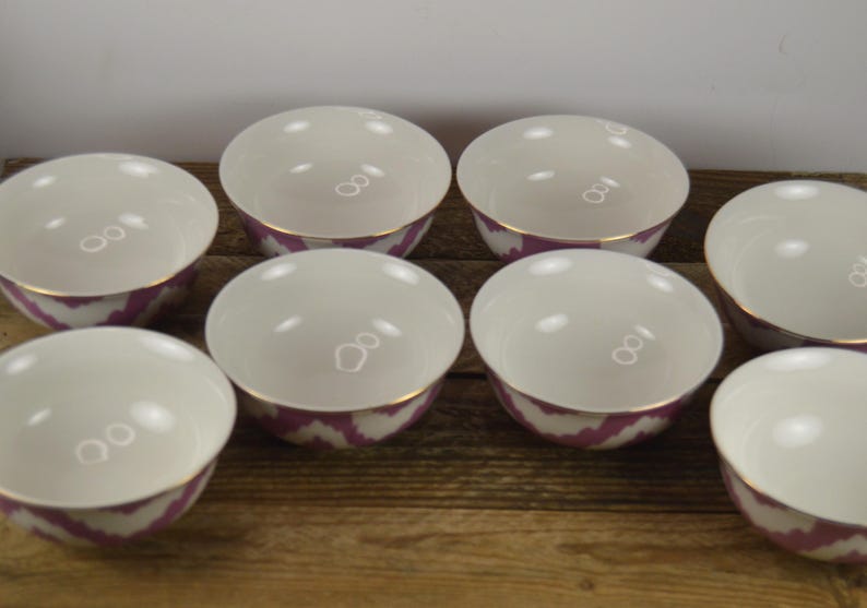 May include: Set of eight white ceramic bowls with a pink and white ikat pattern and gold trim. The bowls are arranged in a circle on a wooden surface.