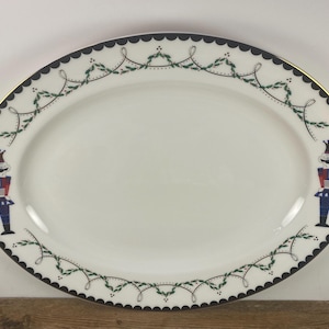 May include: Oval serving platter with a white center, a black scalloped border, and a gold rim. The border features a green and white garland design. Two nutcracker figures in red, blue, and white are on the platter.