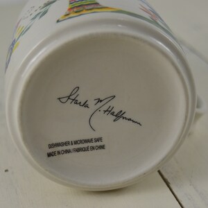 May include: A white ceramic pitcher with a colorful floral design. The bottom of the pitcher features the signature "Starla M. Halfmann" in black script, along with text indicating it is dishwasher and microwave safe. Made in China.