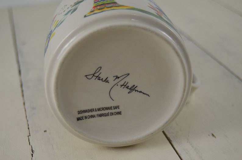 May include: A white ceramic pitcher with a colorful floral design. The bottom of the pitcher features the signature "Starla M. Halfmann" in black script, along with text indicating it is dishwasher and microwave safe. Made in China.