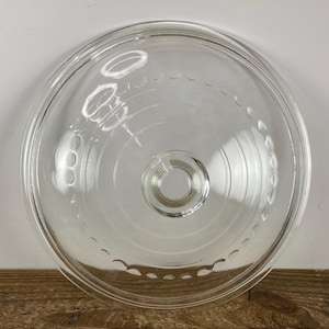 May include: A clear glass lid with a circular design. The lid has a central knob and a series of small, decorative indentations around the edge. The glass is transparent, allowing a view of the surface below. The lid is likely for a pot or pan.