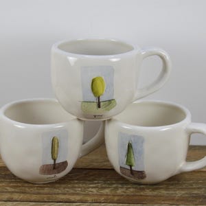 May include: Three white ceramic mugs with tree designs. The mugs have a rounded shape and a handle. The trees are painted in shades of green, yellow, and brown. The mugs are stacked on top of each other.