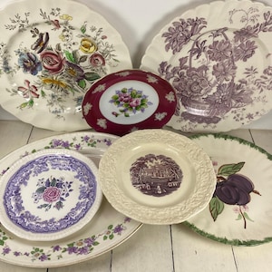May include: A collection of vintage decorative plates with floral and fruit designs. The plates feature a variety of colors, including purple, green, red, and cream. Some plates have scalloped edges, while others have smooth rims. The plates are arranged on a white surface.