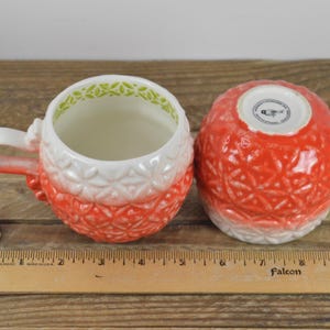 May include: Two red and white ceramic mugs with a textured design. The mugs have a green rim and a white interior. The mugs are sitting on a wooden surface.