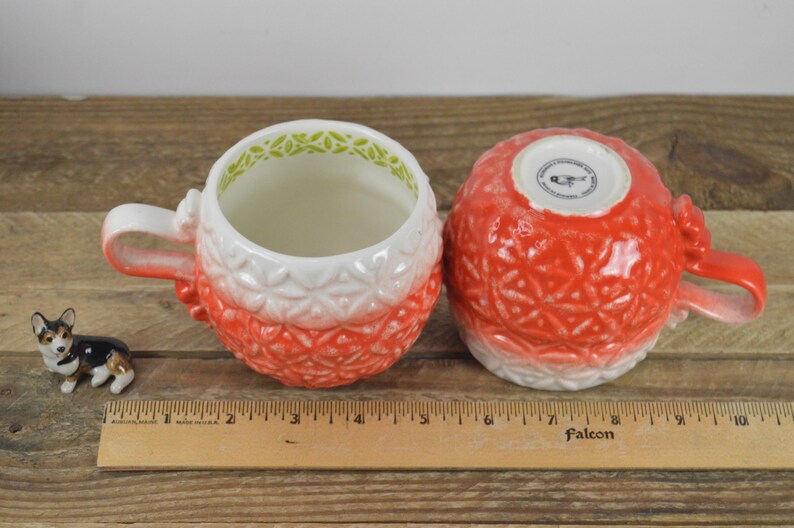 May include: Two red and white ceramic mugs with a textured design. The mugs have a green rim and a white interior. The mugs are sitting on a wooden surface.