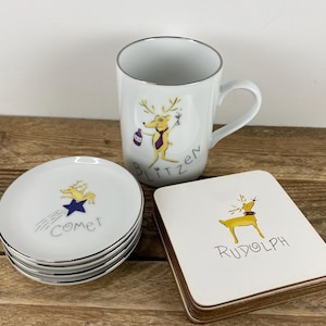 May include: A white ceramic mug, coasters, and small plates with silver trim. The mug features a cartoon reindeer with the name "Blitzen." The plates have "Comet" and a star. The coasters have "Rudolph."