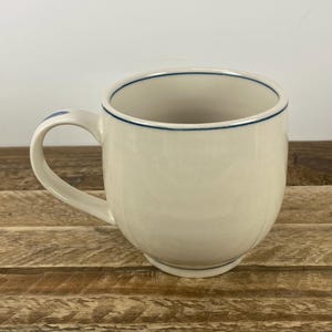 K&ouml;nnte beinhalten: Cremefarbene Keramiktasse mit runder Form und gebogenem Henkel. Der Rand und die Basis des Griffs sind mit einer d&uuml;nnen blauen Linie akzentuiert. Die Tasse steht auf einer Holzoberfl&auml;che.