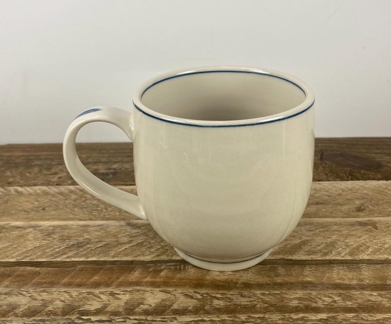 K&ouml;nnte beinhalten: Cremefarbene Keramiktasse mit runder Form und gebogenem Henkel. Der Rand und die Basis des Griffs sind mit einer d&uuml;nnen blauen Linie akzentuiert. Die Tasse steht auf einer Holzoberfl&auml;che.