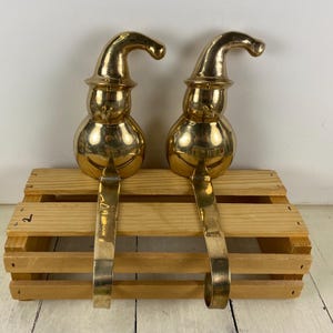 May include: Two brass snowman-shaped Christmas stocking holders. Each holder has a curved hat, round body, and a long, curved hook for hanging stockings. The holders are placed on a wooden crate.