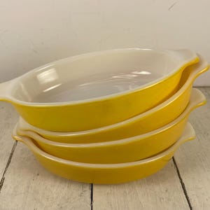 May include: A stack of four oval-shaped baking dishes with bright yellow exteriors and smooth white interiors. Each dish features integrated handles on both sides, creating a uniform appearance for this set of kitchen bakeware.