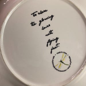 May include: White ceramic plate with a scalloped edge. The plate features black script that reads "To close the glowing hours with flying feet." A dragonfly illustration is also present.
