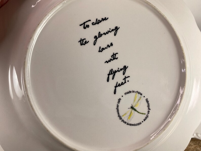 May include: White ceramic plate with a scalloped edge. The plate features black script that reads "To close the glowing hours with flying feet." A dragonfly illustration is also present.