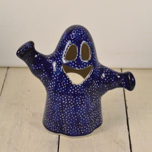 May include: A blue ceramic ghost figurine with white polka dots. The ghost has a smiling face with cut-out eyes and mouth. The ghost is holding out one arm and has a small hole in the top of its head.