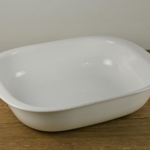 May include: A white, rectangular baking dish with rounded corners. The dish has a smooth, glossy finish and is set against a wooden background. This versatile dish is suitable for baking and serving various dishes.