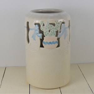 May include: A white ceramic vase with a cutout design of cacti and blue ribbons. The vase has a light pink and blue glaze.