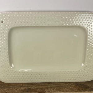 May include: Cream-colored rectangular serving tray with a textured, dotted border. The tray has a smooth, recessed center and a small hole on one side. The tray is made of ceramic or porcelain and is suitable for serving food.