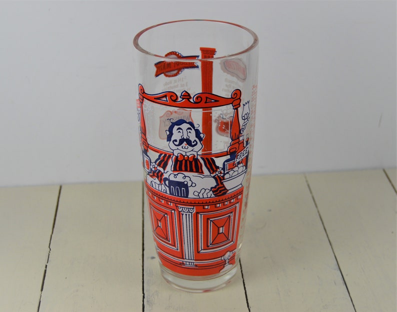 May include: A clear glass with a red and blue illustration of a man with a mustache behind a bar. The glass has measurements for different drinks.