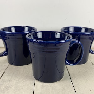 May include: Three matching deep navy blue ceramic mugs with a glossy finish. Each mug features a wide, flared rim, a sturdy C-shaped handle, and a series of three horizontal embossed rings circling the upper portion of the exterior body.