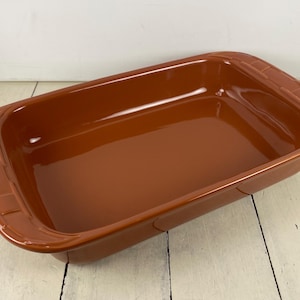 May include: A rectangular, terracotta-colored baking dish with rounded corners and a glossy finish. The dish has slightly raised handles on each end. The interior is smooth and reflective, suggesting it's unused.