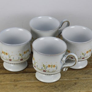 May include: Set of four white ceramic mugs with floral designs. Each mug has a footed base and a handle.