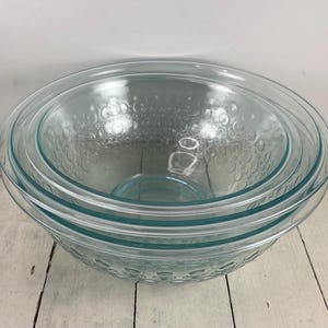 May include: A set of three nested clear glass mixing bowls featuring a textured exterior pattern of raised circular bubbles. The bowls are stacked inside one another, showcasing a smooth interior surface and a consistent light blue-green tint.