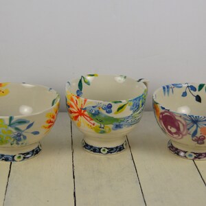 May include: Three ceramic bowls with colourful floral and bird designs. The bowls have a white background with blue, green, yellow, orange, and pink accents. The bowls are on a white wooden surface.