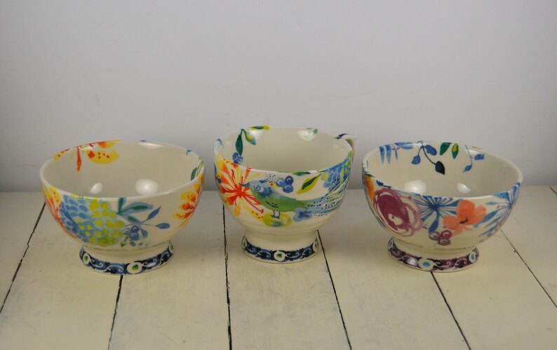May include: Three ceramic bowls with colourful floral and bird designs. The bowls have a white background with blue, green, yellow, orange, and pink accents. The bowls are on a white wooden surface.