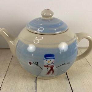 May include: A ceramic teapot with a winter theme. The teapot is painted with a snowman design, featuring a blue hat and scarf. The lid and body have a light blue and white color scheme, with a round handle and spout.