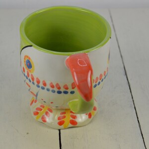 May include: A ceramic mug with a colorful, whimsical design. The mug features a white base with red, blue, yellow, and green accents. The handle is a bright orange color. The mug is shaped like a bird with a beak and feet.