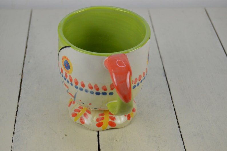 May include: A ceramic mug with a colorful, whimsical design. The mug features a white base with red, blue, yellow, and green accents. The handle is a bright orange color. The mug is shaped like a bird with a beak and feet.
