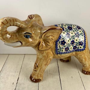 May include: A ceramic elephant figurine with a glossy tan finish and a raised trunk. The elephant features a decorative blanket pattern on its back, adorned with blue flowers, green leaves, and small hearts against a cream-colored background.