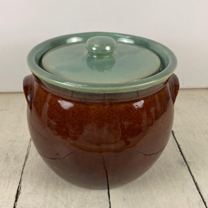 May include: A brown ceramic pot with a lid. The pot has a rounded shape with two small handles on the sides. The lid is a light green color with a small knob on top. The pot is glazed and has a glossy finish.