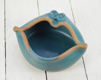 May include: A teal blue ceramic dish shaped like a pair of lips with a brown rim. The dish has two small, round, blue knobs on top.