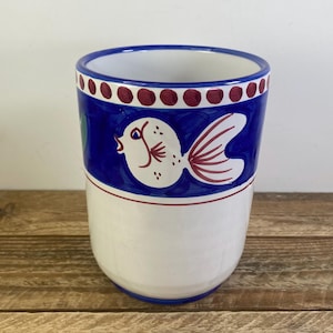 May include: A cylindrical ceramic tumbler featuring a white base and a wide blue band around the top. The blue section is decorated with a white fish illustration with red fins and a border of small red dots along the upper rim of the vessel.