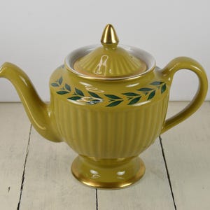 May include: A yellow teapot with a gold trim and green leaf design. The teapot has a lid and a spout.