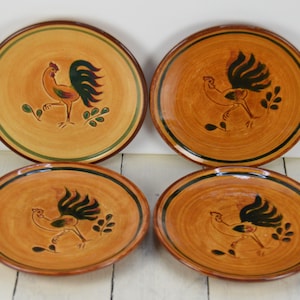 May include: Set of four ceramic plates with a rooster design. The plates are brown with green and black accents. Each plate features a rooster with a green tail and black body.