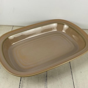 May include: A rectangular serving platter with a rounded edge. The ceramic dish has a light brown interior and a darker brown rim. The platter is on a white wooden surface.