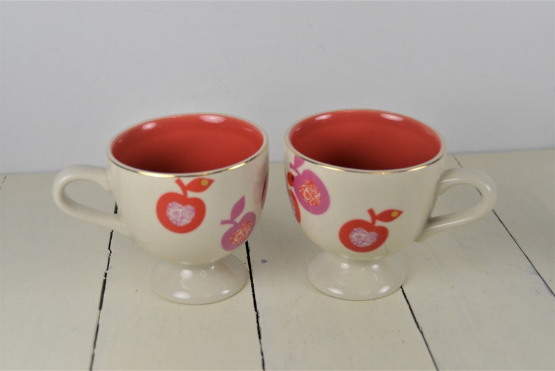 May include: Two white ceramic mugs with a footed base. The mugs have a red interior and are decorated with pink and red apples with gold accents.