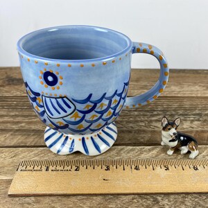 Op de afbeelding: Een lichtblauwe keramische mok met een visontwerp, met blauwe en gele schubben, een ronde handgreep en een voetstuk. Naast de mok staat een klein corgi-beeldje. De mok is ongeveer 10 cm hoog.