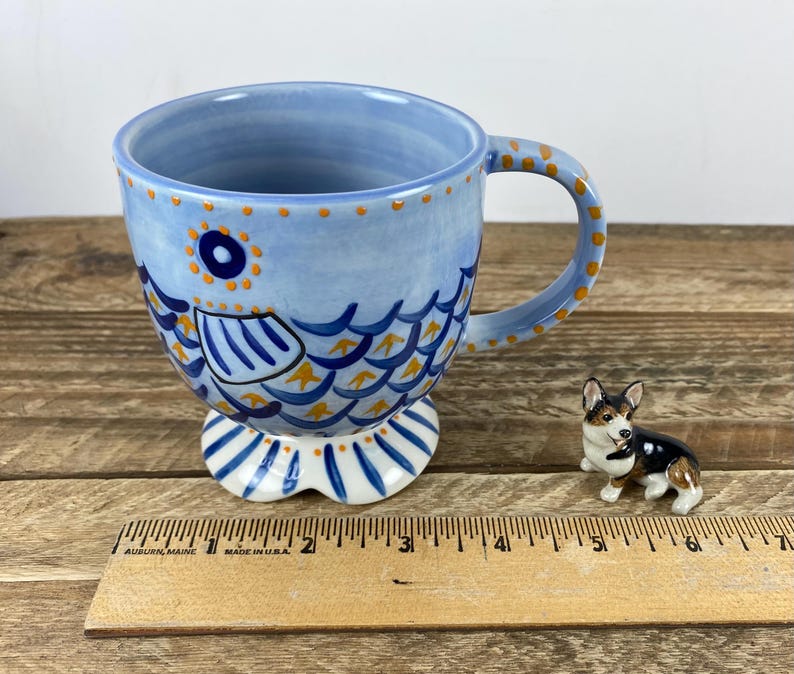 Op de afbeelding: Een lichtblauwe keramische mok met een visontwerp, met blauwe en gele schubben, een ronde handgreep en een voetstuk. Naast de mok staat een klein corgi-beeldje. De mok is ongeveer 10 cm hoog.