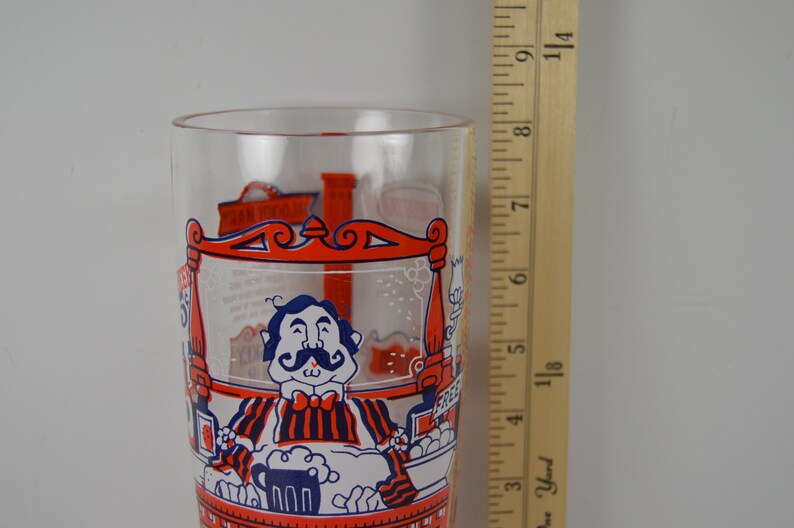 May include: A clear glass with a red, white, and blue illustration of a man with a mustache sitting at a bar with a beer mug. The glass is 7 inches tall.