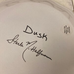 May include: A white ceramic plate with a wavy edge. The plate has the word "Dusk" written in black script, along with a signature. A small sticker on the plate reads "Made in China".