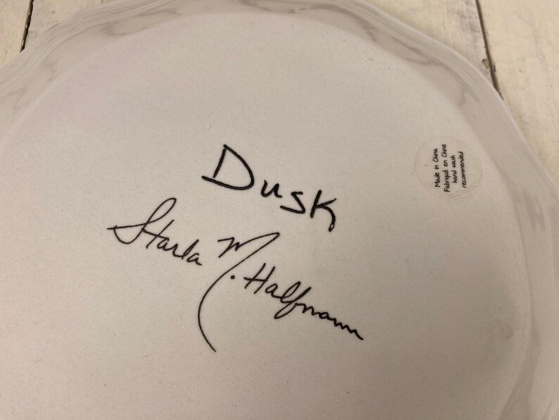 May include: A white ceramic plate with a wavy edge. The plate has the word "Dusk" written in black script, along with a signature. A small sticker on the plate reads "Made in China".
