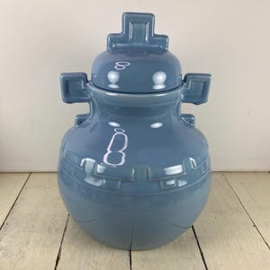 May include: A light blue ceramic jar with a lid. The jar has a rounded body with a geometric design around the middle and square handles on the sides. The lid has a cross-shaped handle. The jar is on a white wooden surface.