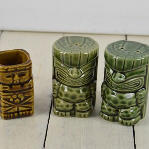 May include: Three ceramic tiki salt and pepper shakers. Two are green with a tiki face design and one is brown with a tiki face design.