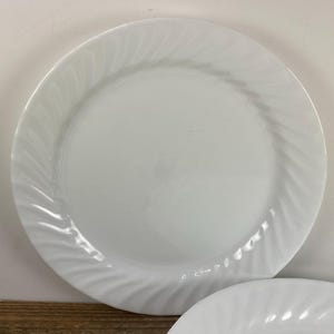 May include: A round white dinner plate featuring a smooth center and a decorative rim with a subtle, raised swirl pattern. The plate has a glossy finish and a clean, uniform white color throughout, with a slightly curved edge design.