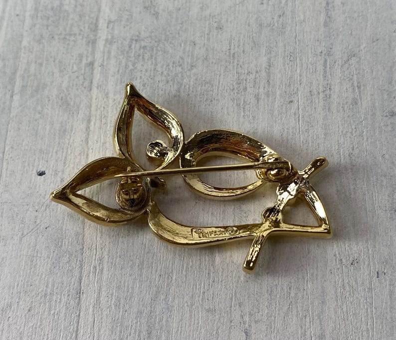 Op de afbeelding: Een goudkleurige broche met een bloemmotief. De broche heeft drie bloemblaadjes en een gebogen, langwerpige vorm. De speld is zichtbaar aan de achterkant.