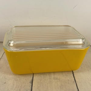 May include: A rectangular yellow glass storage container with a clear glass lid. The lid features a series of parallel horizontal ridges across its surface. The container has smooth, solid yellow sides and a tapered base, providing a simple design.