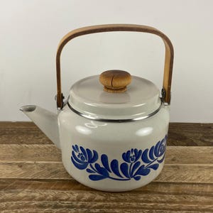 May include: A white enamel teapot with a wooden handle and lid knob. The teapot features a blue floral design around the base. The handle is made of light-colored wood, and the lid knob is a darker wood.